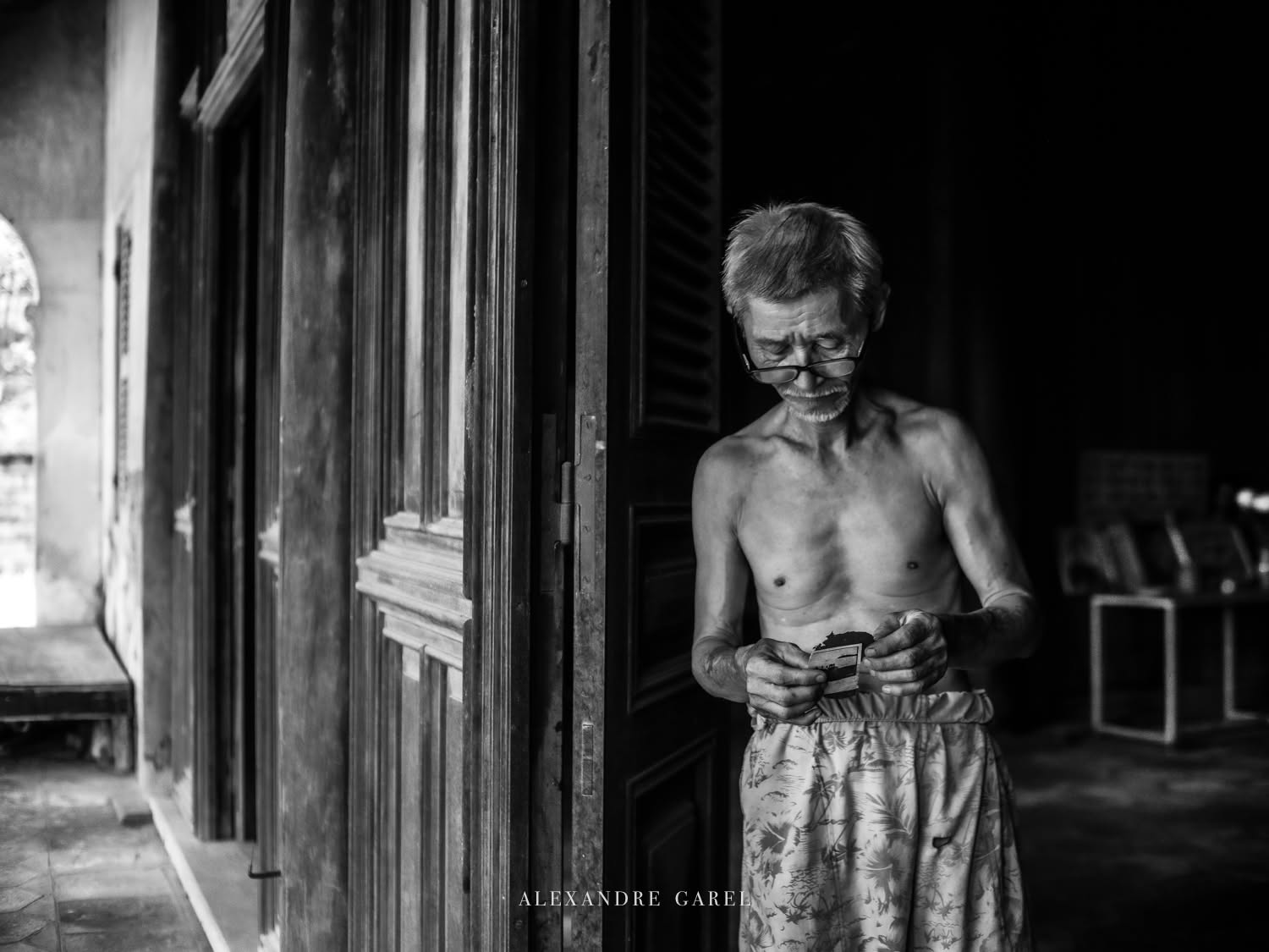 Một người đàn ông lớn tuổi đứng bên cửa gỗ, chăm chú xem tấm ảnh cũ